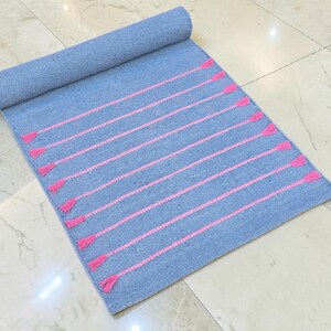 May include: A rolled-up blue rug with pink horizontal stripes and tassels. The rug is made of woven fabric and is partially unrolled, displaying the pink accents. The rug is on a marble tile floor.