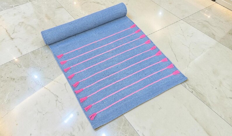 May include: A rolled-up blue rug with pink horizontal stripes and tassels. The rug is made of woven fabric and is partially unrolled, displaying the pink accents. The rug is on a marble tile floor.