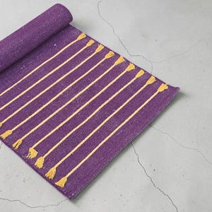 May include: Rolled-up purple yoga mat with yellow stripes and tassels. The mat is partially unrolled, displaying the striped design. The mat is on a gray concrete surface.