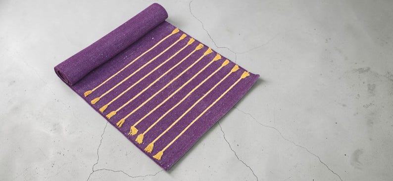 May include: Rolled-up purple yoga mat with yellow stripes and tassels. The mat is partially unrolled, displaying the striped design. The mat is on a gray concrete surface.
