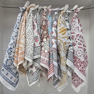 May include: A collection of colorful, patterned cotton scarves hanging from a metal rod. The scarves feature various designs, including floral, geometric, and paisley patterns, in shades of blue, brown, white, and red. The scarves are knotted at the top.