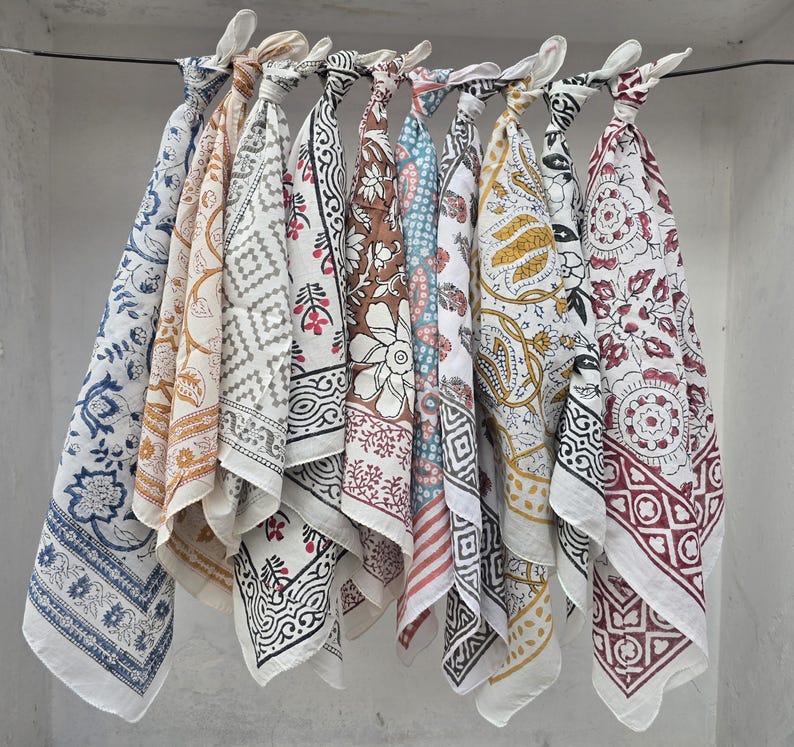 May include: A collection of colorful, patterned cotton scarves hanging from a metal rod. The scarves feature various designs, including floral, geometric, and paisley patterns, in shades of blue, brown, white, and red. The scarves are knotted at the top.