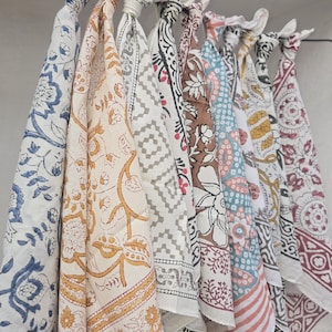 May include: A collection of colorful, patterned fabric cloths hanging from a rod. The cloths feature various designs, including floral, geometric, and paisley patterns in shades of blue, yellow, brown, and red. Each cloth is knotted at the top.