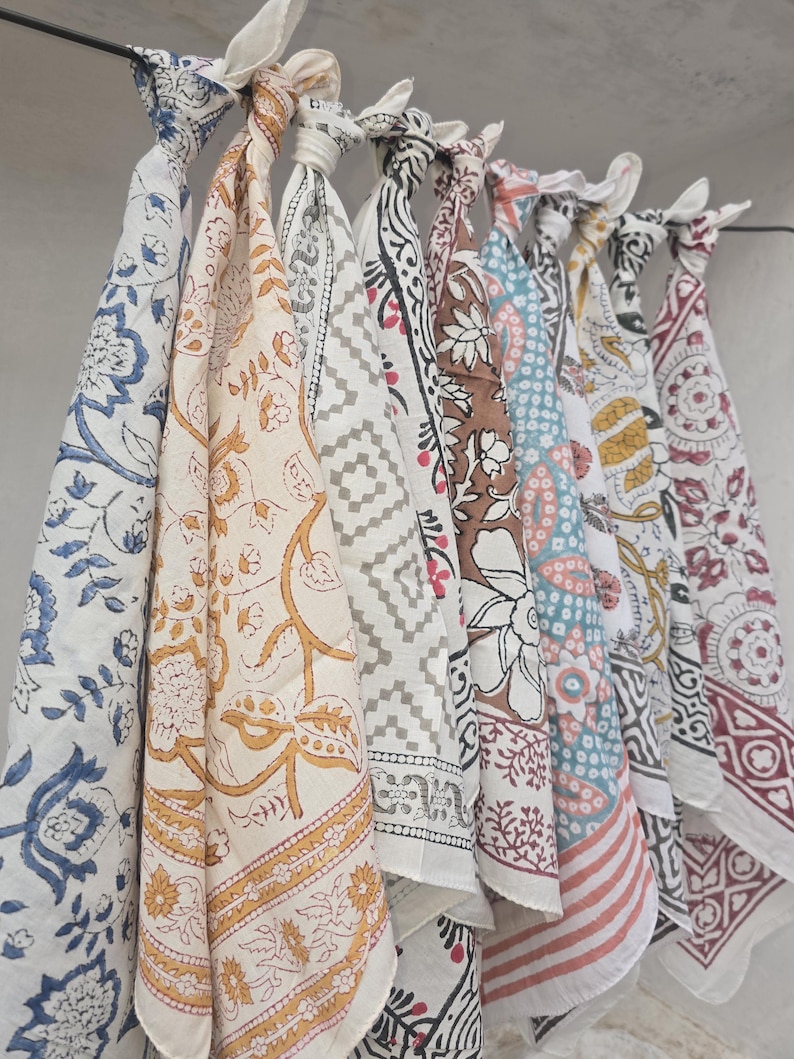 May include: A collection of colorful, patterned fabric cloths hanging from a rod. The cloths feature various designs, including floral, geometric, and paisley patterns in shades of blue, yellow, brown, and red. Each cloth is knotted at the top.