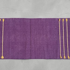 May include: A rectangular purple table runner with vertical gold tassels along the edges. The runner is made of a textured fabric. The tassels are evenly spaced and add a decorative element to the runner.