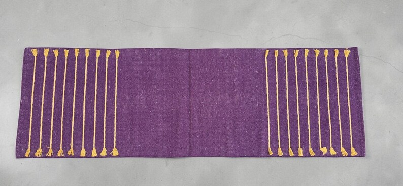 May include: A rectangular purple table runner with vertical gold tassels along the edges. The runner is made of a textured fabric. The tassels are evenly spaced and add a decorative element to the runner.