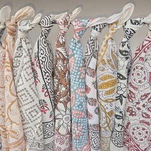 May include: A collection of patterned scarves in various colors, including blue, brown, and white, are displayed hanging from a metal rod. Each scarf features a unique design, such as floral or geometric patterns, and is knotted at the top.