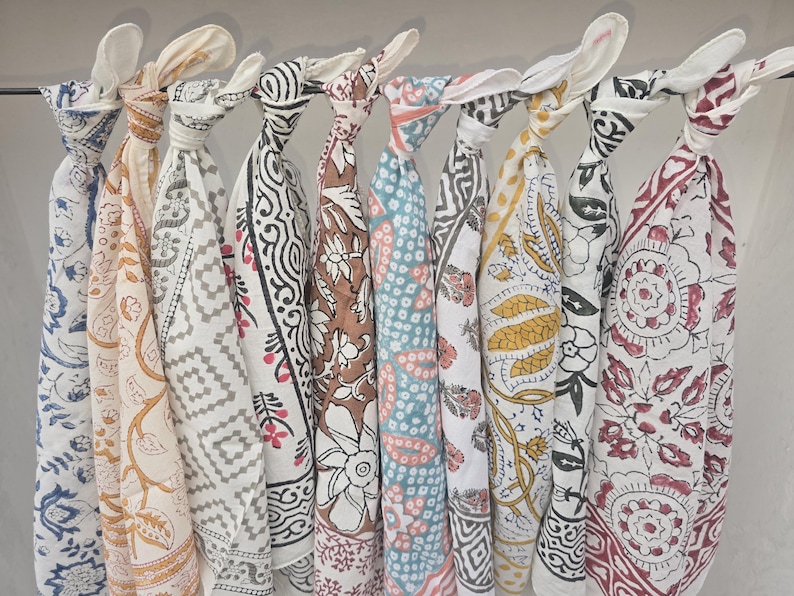 May include: A collection of patterned scarves in various colors, including blue, brown, and white, are displayed hanging from a metal rod. Each scarf features a unique design, such as floral or geometric patterns, and is knotted at the top.