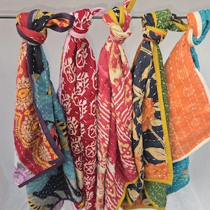 Op de afbeelding: Een verzameling kleurrijke, bedrukte sjaals die aan een metalen stang hangen. De sjaals hebben verschillende ontwerpen, waaronder bloemen- en geometrische patronen, in tinten rood, blauw, oranje en geel. Elke sjaal is bovenaan geknoopt.