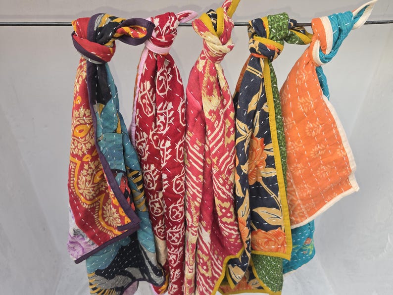 Op de afbeelding: Een verzameling kleurrijke, bedrukte sjaals die aan een metalen stang hangen. De sjaals hebben verschillende ontwerpen, waaronder bloemen- en geometrische patronen, in tinten rood, blauw, oranje en geel. Elke sjaal is bovenaan geknoopt.