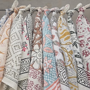 May include: A collection of colorful, patterned bandanas hanging from a rod. The bandanas feature various designs, including floral, geometric, and paisley patterns in shades of blue, yellow, brown, and red. Each bandana is knotted at the top.