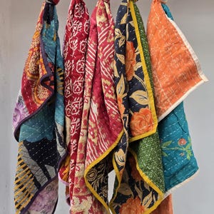 Op de afbeelding: Een verzameling kleurrijke, bedrukte stoffen sjaals die aan een stang hangen. De sjaals hebben verschillende ontwerpen, waaronder bloemen- en geometrische patronen, in tinten rood, blauw, oranje en groen. Elke sjaal is bovenaan geknoopt.