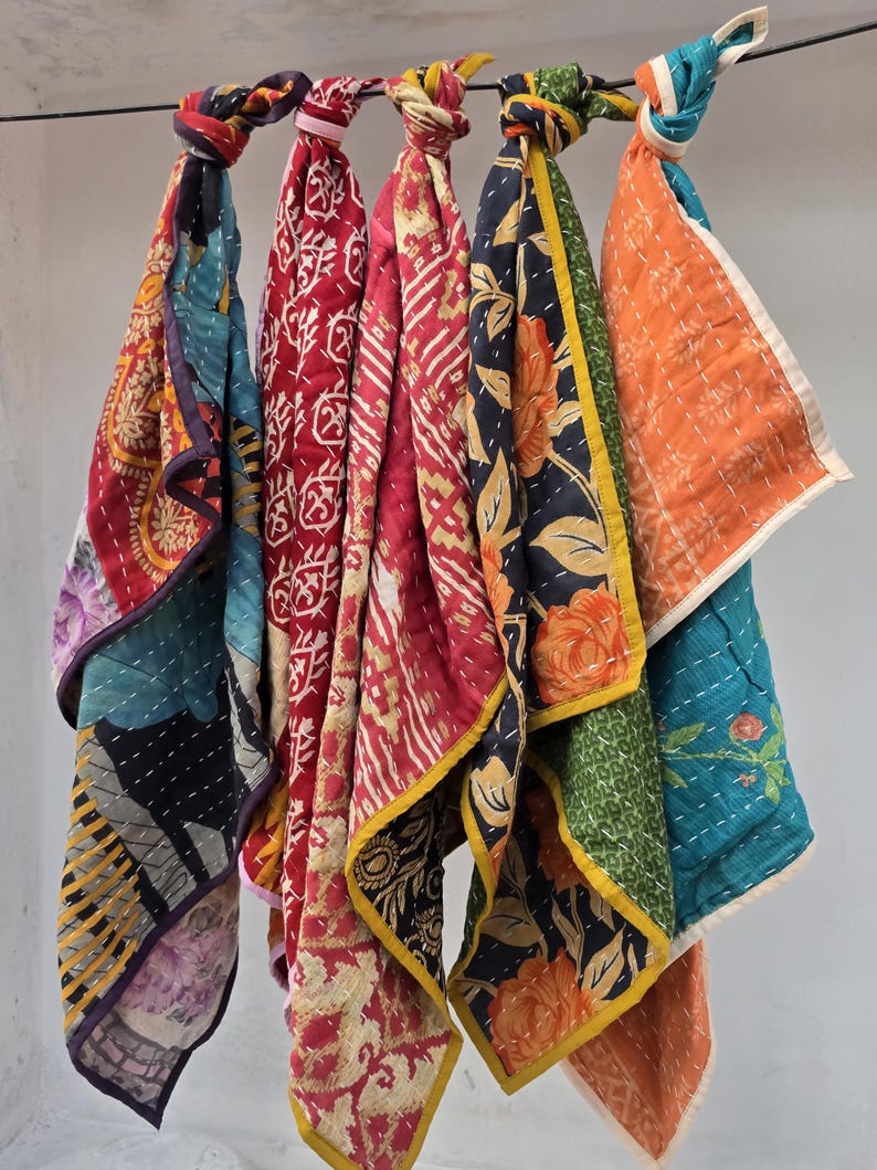 Op de afbeelding: Een verzameling kleurrijke, bedrukte stoffen sjaals die aan een stang hangen. De sjaals hebben verschillende ontwerpen, waaronder bloemen- en geometrische patronen, in tinten rood, blauw, oranje en groen. Elke sjaal is bovenaan geknoopt.