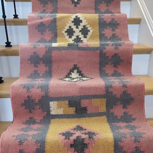May include: A stair runner rug with a geometric pattern in shades of pink, grey, and yellow. The rug is installed on wooden stairs and features a repeating design of diamonds and floral shapes.