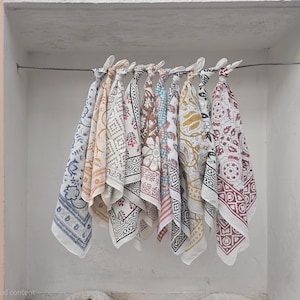 May include: A collection of colorful, patterned cotton scarves hanging on a wire. The scarves feature various designs, including floral, geometric, and paisley patterns in shades of blue, red, yellow, and brown. The scarves are displayed against a white wall.