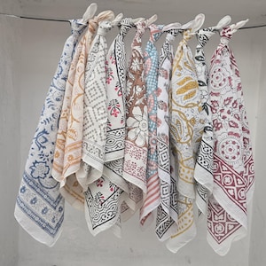 May include: A collection of patterned cotton bandanas in various colors, including blue, yellow, brown, and red, hanging from a metal rod. Each bandana features a unique floral or geometric design, with white borders.