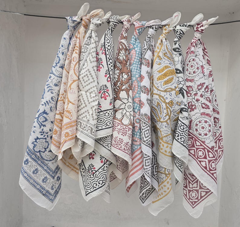 May include: A collection of patterned cotton bandanas in various colors, including blue, yellow, brown, and red, hanging from a metal rod. Each bandana features a unique floral or geometric design, with white borders.