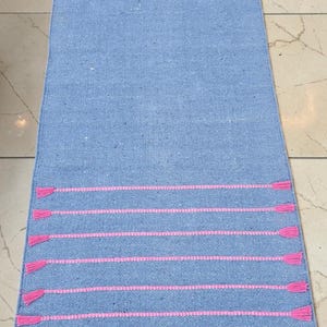 May include: A rectangular, light blue rug with horizontal pink stripes and pink tassels at each end. The rug is made of woven material and is displayed on a tiled floor. The rug is a home decor item.
