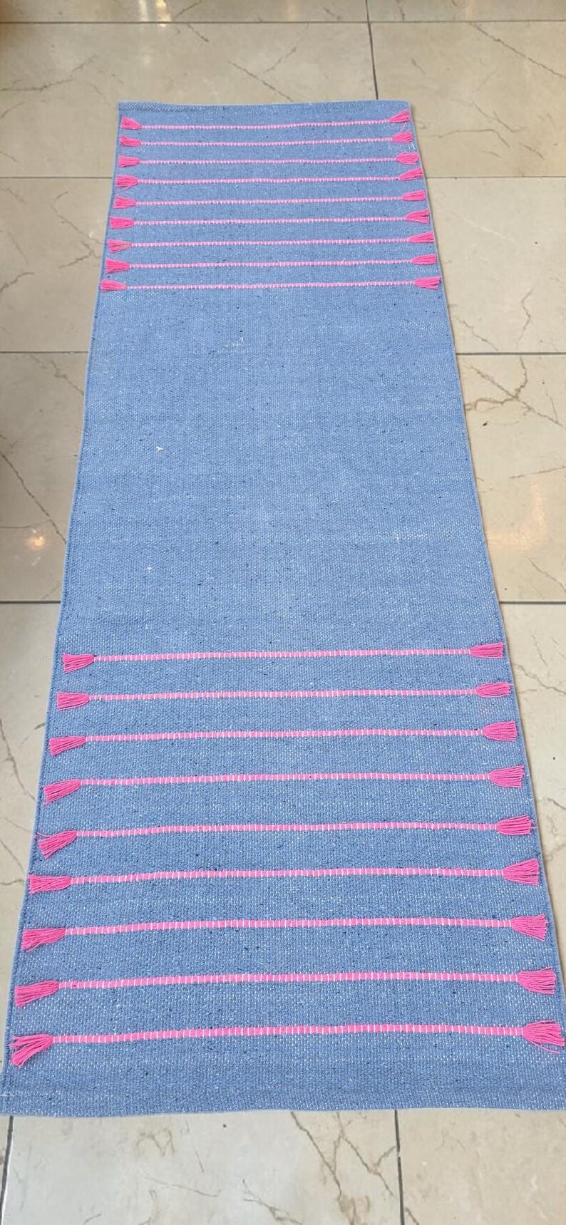 May include: A rectangular, light blue rug with horizontal pink stripes and pink tassels at each end. The rug is made of woven material and is displayed on a tiled floor. The rug is a home decor item.