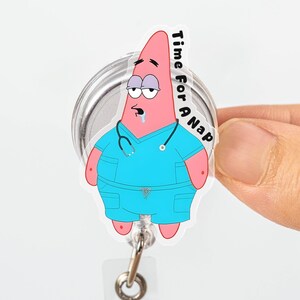 May include: A pink Patrick Star badge reel with the text "Time For A Nap." The cartoon character is wearing blue scrubs and a stethoscope. The badge reel has a clear plastic retractable cord and a metal clip.