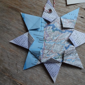 May include: A handmade star-shaped ornament crafted from a map, featuring a detailed geographical design. The star is folded with multiple points and has a small metal eyelet for hanging. The ornament is placed on a wooden surface.