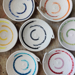 Puede incluir: Conjunto de siete cuencos redondos de cuerda trenzada con detalles de tela de colores. Los cuencos son blancos con una variedad de colores, incluyendo azul, amarillo, verde, naranja y rojo.