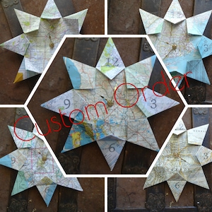 Puede incluir: Un reloj en forma de estrella hecho con mapas antiguos plegados. La esfera del reloj presenta un mecanismo de reloj dorado con manecillas y números negros. Las palabras "Custom Order" están escritas en rojo en la imagen.