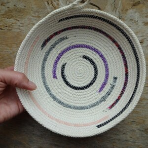 May include: A white woven bowl with a circular design featuring black, gray, purple, and pink rope. The bowl is handmade and has a unique, modern look.