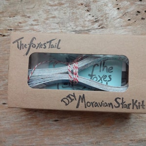 May include: A rectangular brown cardboard box with a clear window displaying paper strips tied with red and white twine. The box features the handwritten text The Foxes Tail and DIY Moravian Star Kit in black ink on the front surface.