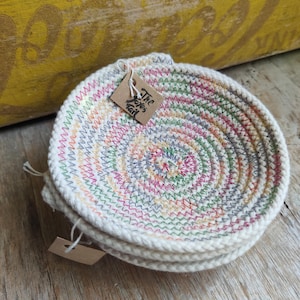 May include: A stack of three handmade, circular coasters. Each coaster is made of off-white rope, with colorful stitching in a spiral pattern. The coasters have a tag that reads "The Eyes Tail". The coasters are on a wooden surface.