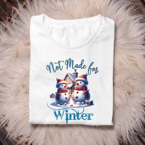 May include: White t-shirt featuring a winter-themed graphic. The design includes two snowmen wearing red hats and scarves, with a house in the background. The text "Not Made for Winter" is displayed above and below the snowmen.