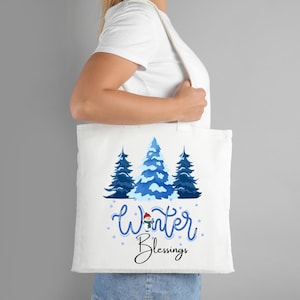 May include: White tote bag featuring a winter scene with three blue pine trees and the words "Winter Blessings" in blue script. A snowman graphic is included in the design. The bag is carried by a person.