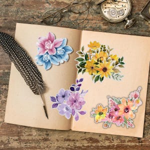 May include: An open book displays four floral sticker designs. The stickers feature watercolor-style flowers in pink, blue, yellow, purple, and orange. A black and white feather and a pair of glasses are on the left side of the book, and a pocket watch is on the right.