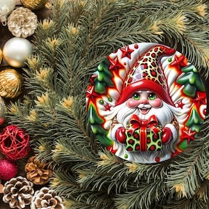 May include: A festive Christmas ornament featuring a gnome design. The gnome wears a red hat with a leopard print pattern and holds a green gift box. The ornament is surrounded by pine needles, red and gold ornaments, and pine cones.