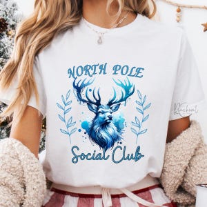 May include: White t-shirt featuring a blue watercolor design of a deer with large antlers. The text "North Pole Social Club" is arched above and below the deer illustration in a script font. The design is surrounded by blue leaves.