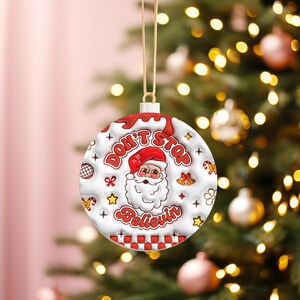 May include: A white Christmas ornament with a red and white Santa Claus design and the words "Don't Stop Believin'". The ornament is round and has a red and white checkered border. It is hanging from a gold string.