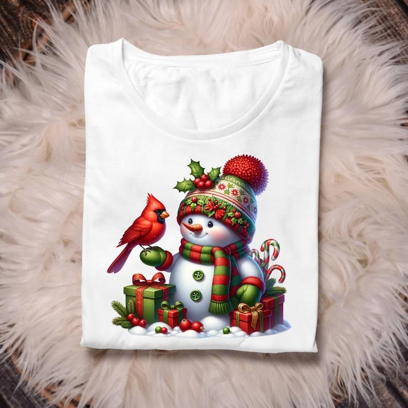 Puede incluir: Camiseta blanca con un dise&ntilde;o de mu&ntilde;eco de nieve festivo. El mu&ntilde;eco de nieve lleva un gorro, bufanda y guantes, y est&aacute; acompa&ntilde;ado por un cardenal rojo y regalos envueltos. El dise&ntilde;o incluye bastones de caramelo y bayas de acebo.