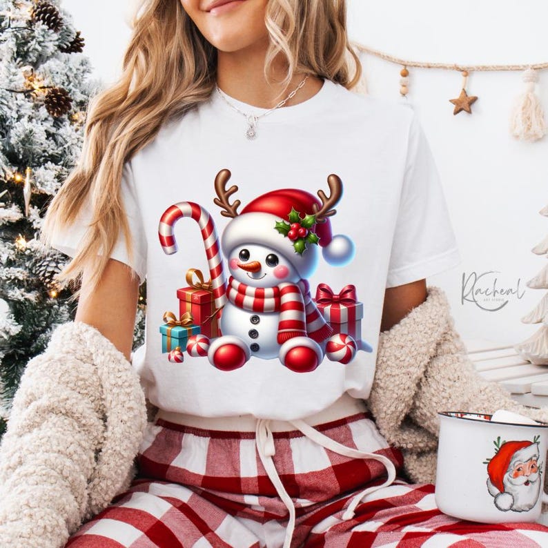 Puede incluir: Camiseta blanca con un dise&ntilde;o de mu&ntilde;eco de nieve navide&ntilde;o. El mu&ntilde;eco de nieve lleva una bufanda y un gorro a rayas rojas y blancas con cuernos, y sostiene un bast&oacute;n de caramelo. Los regalos y las bayas de acebo completan el tema. La camiseta se combina con pantalones a cuadros rojos y blancos.