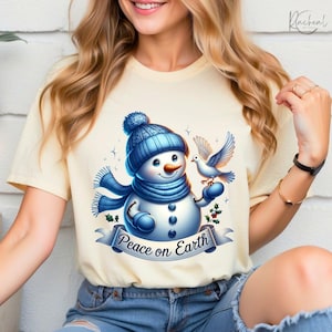 Puede incluir: Camiseta color crema con un alegre mu&ntilde;eco de nieve con gorro y bufanda azules, sosteniendo una paloma. El mu&ntilde;eco de nieve tiene una nariz de zanahoria y ojos de bot&oacute;n. Una pancarta debajo dice "Peace on Earth".