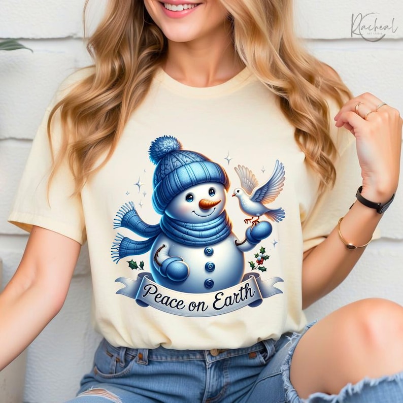 Puede incluir: Camiseta color crema con un alegre mu&ntilde;eco de nieve con gorro y bufanda azules, sosteniendo una paloma. El mu&ntilde;eco de nieve tiene una nariz de zanahoria y ojos de bot&oacute;n. Una pancarta debajo dice "Peace on Earth".