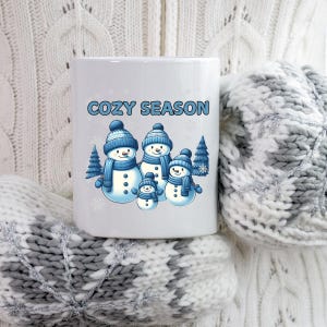 May include: White ceramic mug featuring a family of snowmen wearing blue hats and scarves, with the text "COZY SEASON" in blue. The mug is surrounded by a white and gray knitted sweater and mittens.