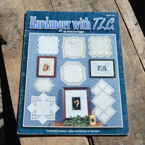 May include: A book titled "Hardanger with T.L.C." by Terry Lee Capps. The cover features a blue background with various white embroidered pieces and framed photos. The book is compiled by Susan L. Meier and Rosalyn K. Watnemo.