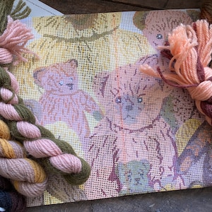 May include: A needlepoint canvas with a design of three koala bears in a forest setting. The canvas is surrounded by skeins of yarn in various shades of brown, pink, and yellow.