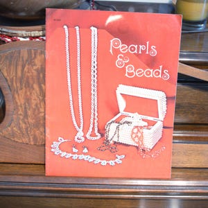 May include: A red brochure titled "Pearls & Beads" features illustrations of white beaded necklaces, earrings, and a jewelry box filled with necklaces. The jewelry box is open, displaying various beaded jewelry.