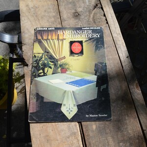 May include: A vintage book titled "Hardanger Embroidery: An Introduction" by Marion Scoular. The cover features a table setting with a white tablecloth and embroidered details, along with a framed artwork and plants. The book is on a wooden surface.