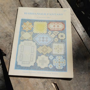 May include: A vintage book titled "Hardanger Fantasy" by Sara Lee Allen. The cover features various cross-stitch patterns in yellow, blue, and white. The book is on a weathered wooden surface.