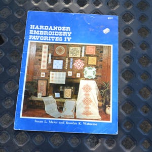 May include: A vintage book titled "Hardanger Embroidery Favorites IV" with a blue cover. The cover features a photo of various embroidered pieces displayed on a brick fireplace. The book is slightly worn.