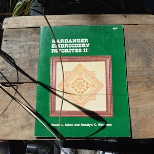 May include: A green book titled "Hardanger Embroidery Favorites II" with a framed embroidery design on the cover. The design is a geometric pattern in shades of orange and cream. The book is on a wooden surface.