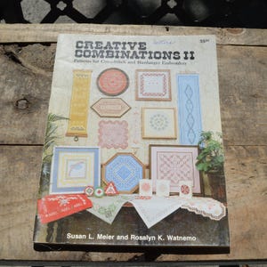 May include: A book titled "CREATIVE COMBINATIONS II" with patterns for cross-stitch and Hardanger embroidery. The cover features various framed embroidery designs in shades of blue, pink, yellow, and red. The book is on a wooden surface.
