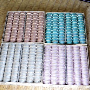 May include: Four square cardboard boxes containing rows of small, round, thread-wrapped bobbins. The bobbins are arranged in columns and come in four distinct colors: light brown, teal, off-white, and pale lavender, each secured with a thin band.